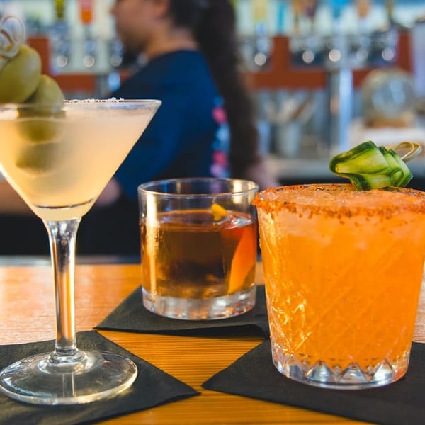 Three cocktails, a martini, an old fashioned, and an orange drink, on a wooden bar.