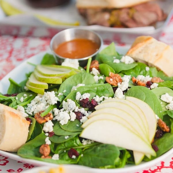 Spinach salad with sliced pears, blue cheese, cranberries, pecans, and dressing on a red patterned tablecloth.