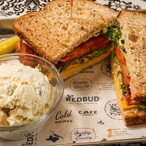 A Redbud Cafe sandwich with potato salad and a pickle on a branded tray liner.