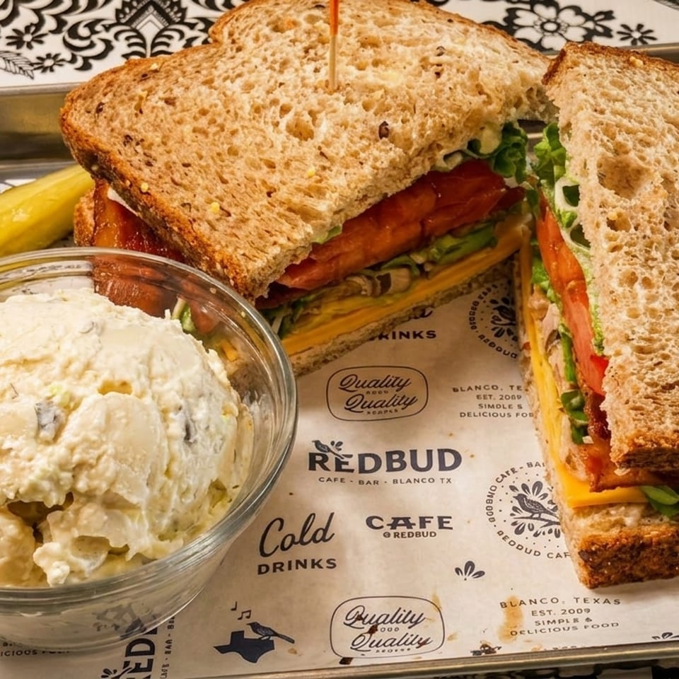 A Redbud Cafe sandwich with potato salad and a pickle on a branded tray liner.