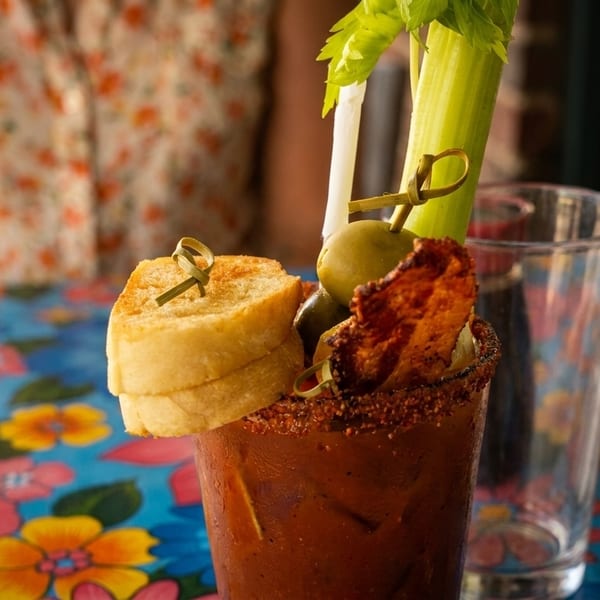 A Bloody Mary cocktail garnished with celery, olives, bacon, and toast on a colorful floral tablecloth.