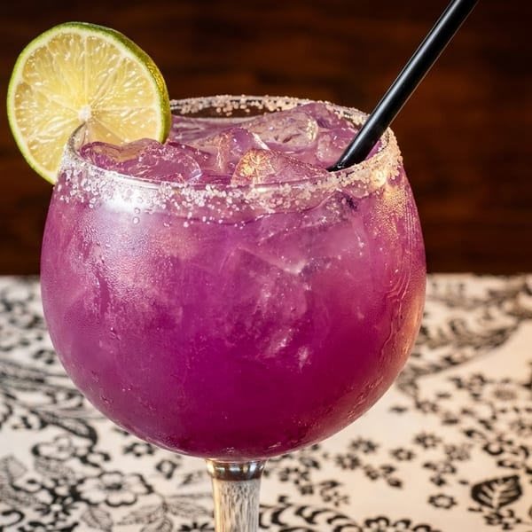 A vibrant purple margarita with a salted rim, lime wedge, and black straw on a patterned tablecloth.